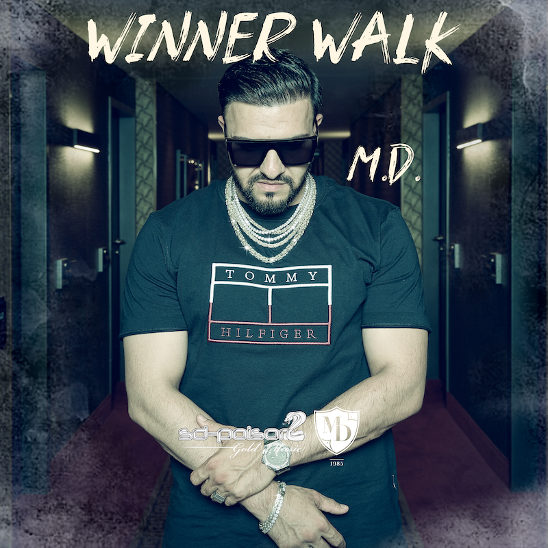 King M.D. steht im Gang eines Luxushotels. Er trägt eine Sonnenbrille, T-Shirt und eine grosse Kette. Es ist das Song Cover vom Lied Winner Walk.